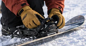 Gloved hands working the ratchet and strap of a black snowboard binding on a board resting in the snow
