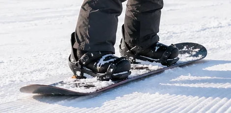 Close-up of snowboard boots in black bindings on a groomed slope, showing the board surface used to butter on a snowboard