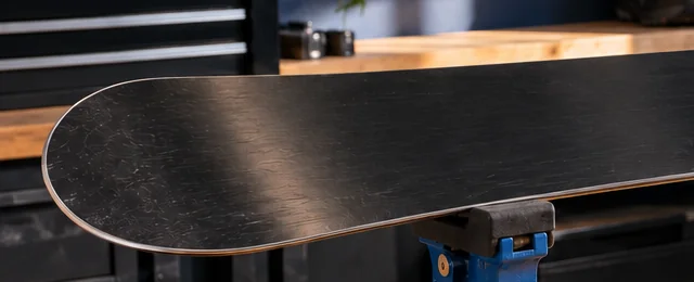 Snowboard clamped base-up in a blue vise on a workbench, with the steel edge visible along the base, ready for rust removal